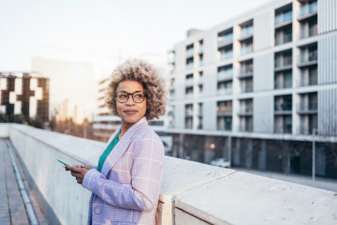 Cheerful professional black woman with blond hair and trendy eyeglasses standing in the city looking away at sunset