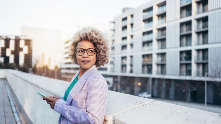 Cheerful professional black woman with blond hair and trendy eyeglasses standing in the city looking away at sunset