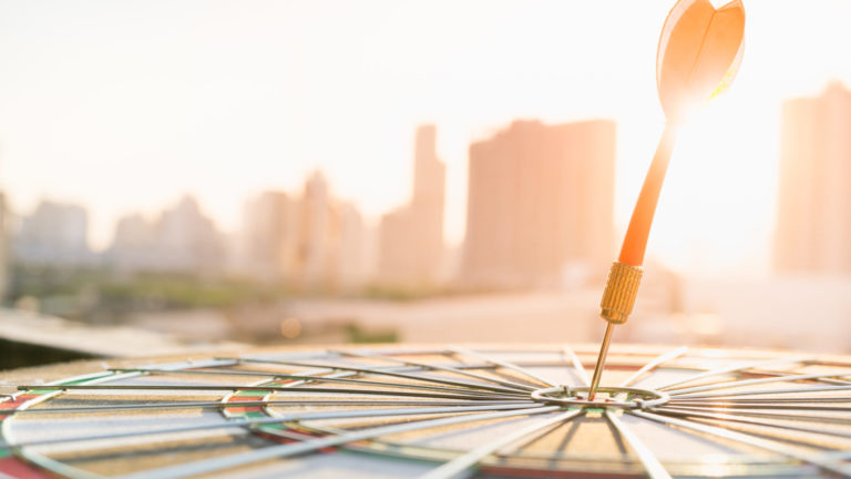 Red dart arrow hitting in the target center of dartboard with modern city and sunset background. Target business, achieve and victory concept .