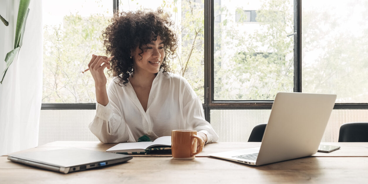 Smiling, happy, Young beautiful african american woman studying at home with laptop. Taking notes in notebook. Bright spacious living room.