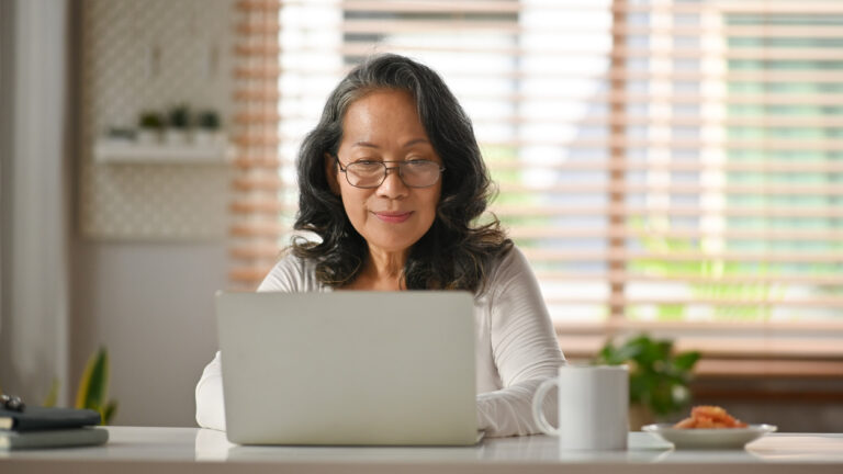 Contemporary senior female in glasses watching webinar or checking email on laptop computer at home.